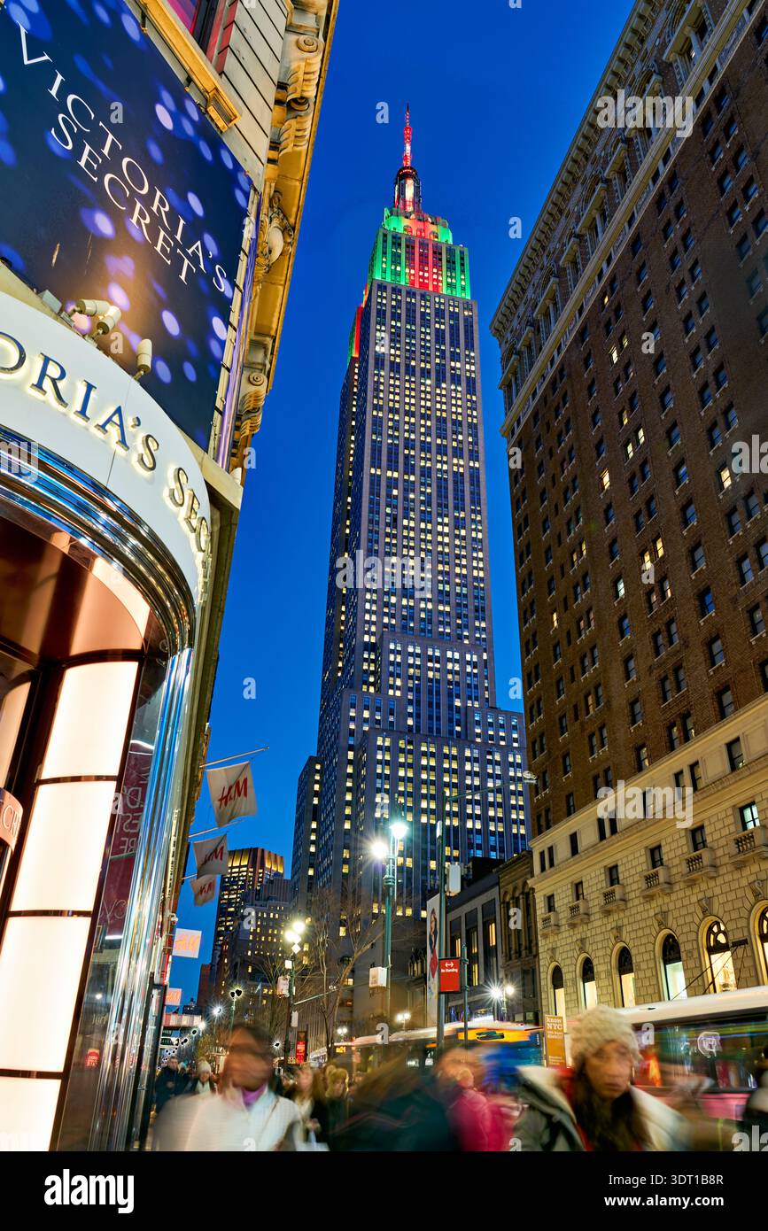 Die Menschen laufen auf der Straße in der Nähe des hell beleuchteten Empire State Building in Manhattan, New York. Die Szene zeigt das Leben und die Lichter der Stadt. Stockfoto
