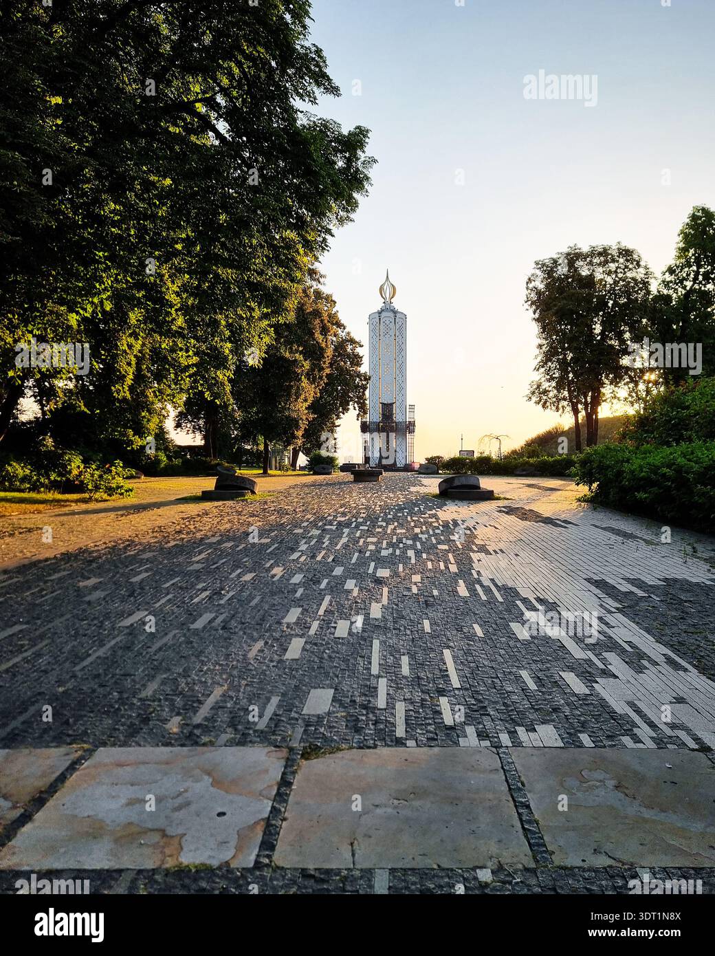 Holodomor-Denkmal in Kiew bei Sonnenaufgang. Der hohe, weiße Turm mit volkstümlichen Mustern, der bei Sonnenaufgang von üppigen grünen Bäumen umgeben ist. Stockfoto