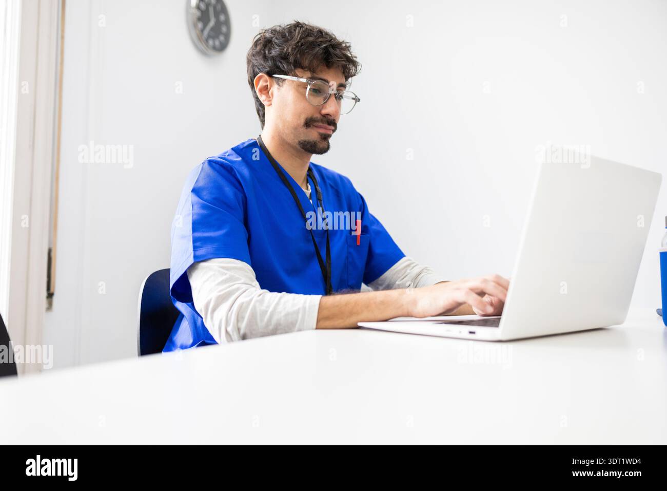 Ein Arzt, der blaue Peelings trägt, konzentriert sich auf die Verwendung eines Laptops an einem Schreibtisch in einem modernen Büro. Die professionelle Umgebung wird durch eine Wanduhr A ergänzt Stockfoto