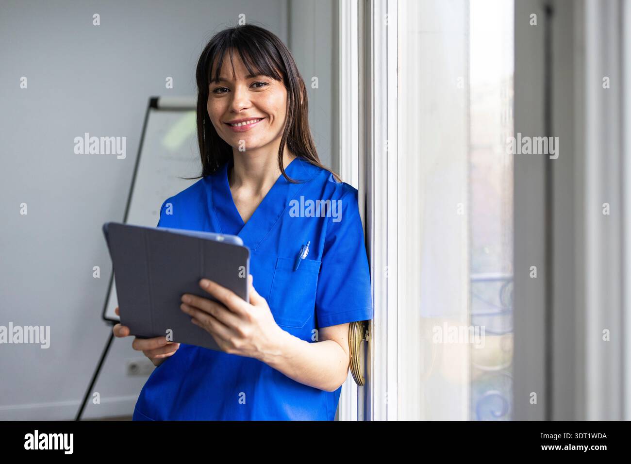 Eine Ärztin in blauem Scrubs hält ein digitales Tablet und lächelt dabei warm in die Kamera. Sie steht an einem Fenster, projiziert Professionalität und ein Juwel Stockfoto