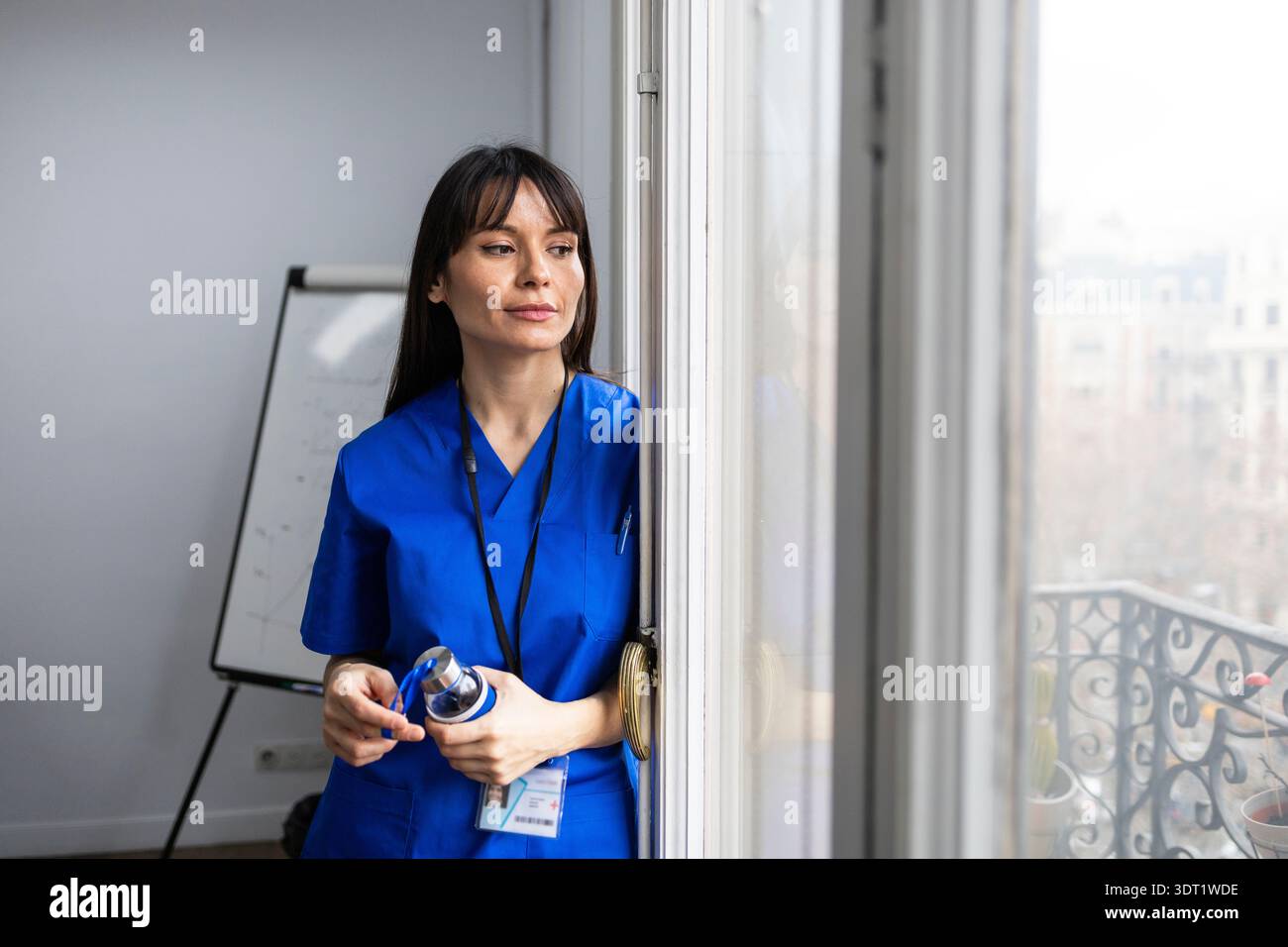Ein Doktor in blauem Gewand steht an einem Fenster und blickt nachdenklich nach draußen. Sie hält ein Schlüsselband und eine Wasserflasche, was für Engagement und Bereitschaft steht Stockfoto