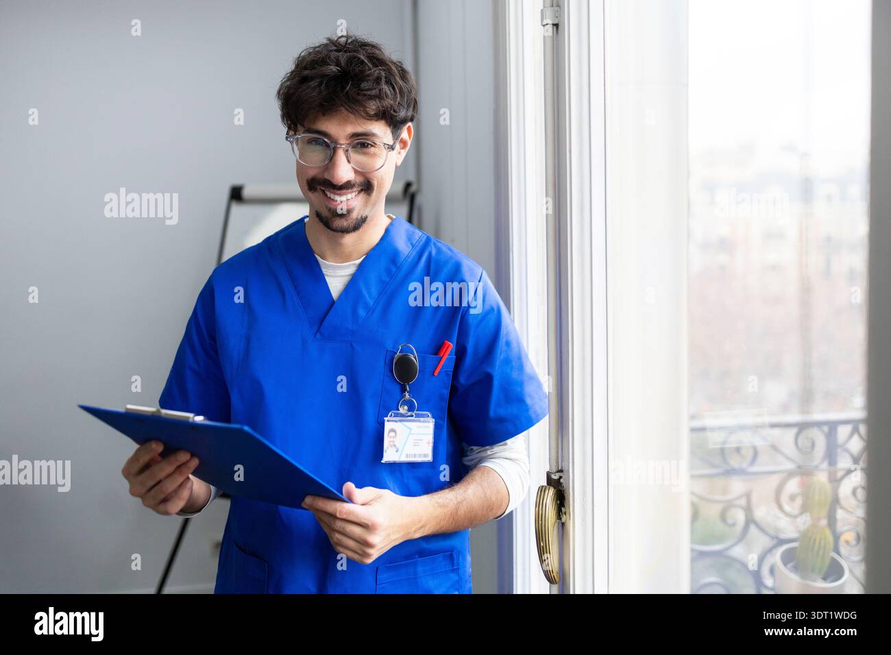 Ein männlicher Arzt, der blaue Peelings trägt, lächelt selbstbewusst, während er ein Klemmbrett an einem hellen Fenster hält. Er steht in einem modernen medizinischen Büro, verkörpert Profi Stockfoto