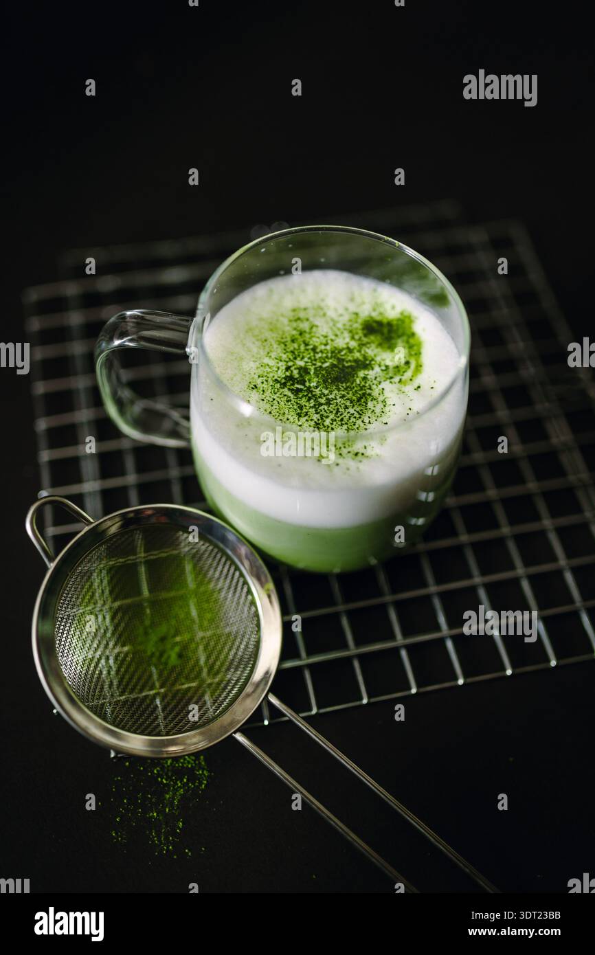 Ein Glasbecher gefüllt mit leuchtend grünem Matcha Latte, garniert mit aufgeschäumter Milch und bestreut mit Matcha-Puderpulver. Die Szene enthält ein feinmaschiges Sieb Stockfoto