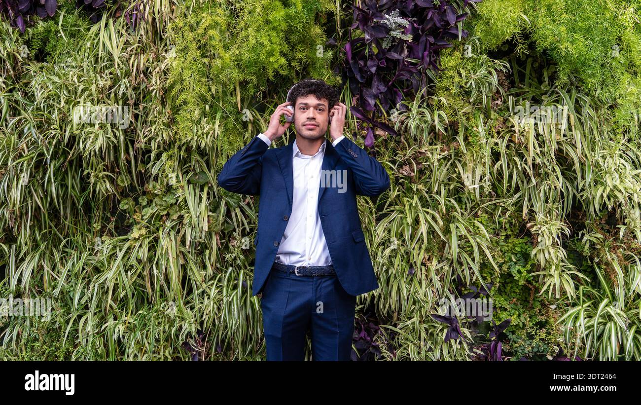 Ein Mann in einem blauen Anzug steht vor einer lebendigen vertikalen Gartenwand mit Kopfhörern, umgeben von üppigen grünen Pflanzen und lila Blättern, kreieren Stockfoto