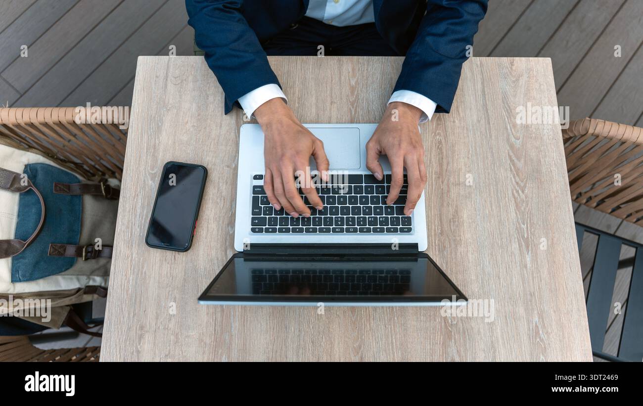 Ein Profi, der einen Anzug trägt, sitzt auf einem Laptop an einem Schreibtisch aus Holz im Freien. In der Nähe befinden sich ein Mobiltelefon und ein Rucksack, um die Geschäftsumgebung zu verbessern. Stockfoto
