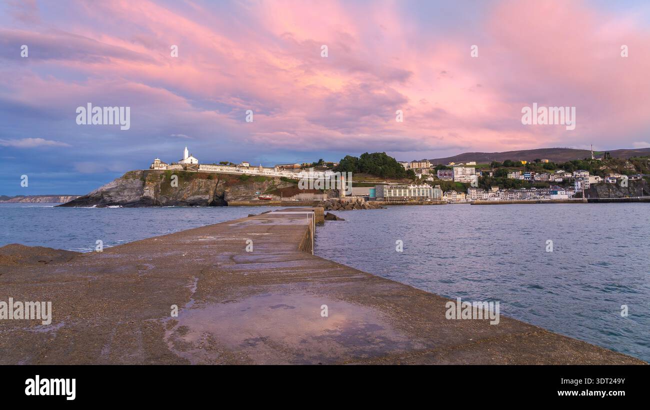 Faszinierendes abendliches Panorama des Hafens von Luarca, das einen atemberaubenden Sonnenuntergang mit rosa und lila Tönen offenbart. Das ruhige Wasser und die malerische Küste schaffen einen Stockfoto