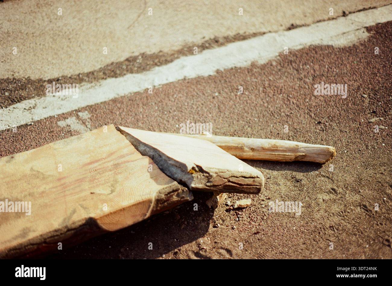 Gebrochene Holzdiele, die bei hellem Sonnenlicht auf einer Asphaltoberfläche liegt, minimale Zusammensetzung mit rauen Texturen und Schatten. Stockfoto