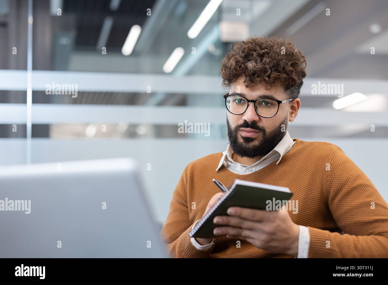 Junger Mann mit lockigem Haar und Bart, der eine Brille trägt, konzentrierte sich auf den Laptop, während er in einem modernen Coworking-Büro Notizen in ein Notizbuch schreibt, plant und rekonstruiert Stockfoto