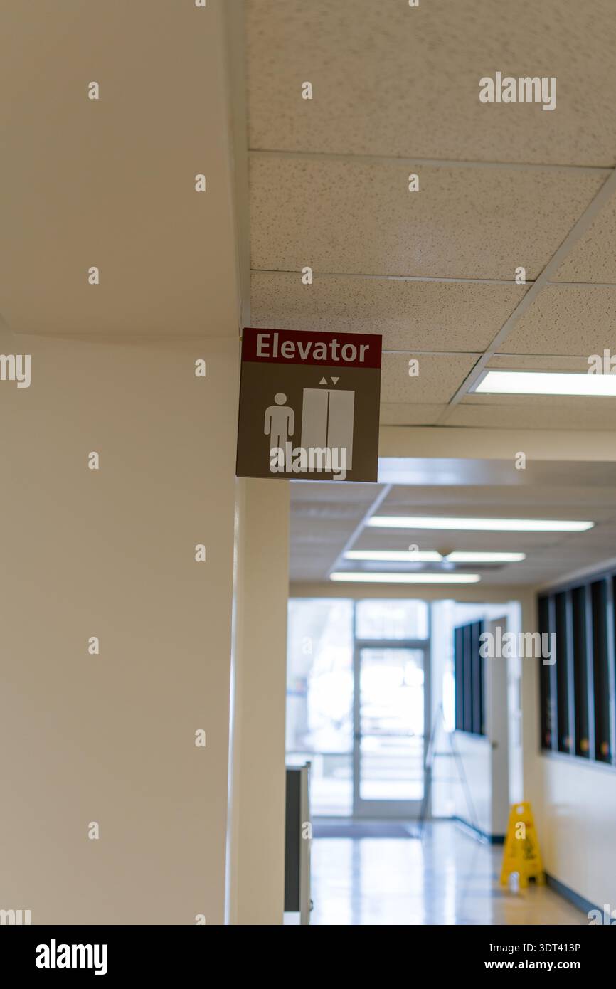Eine vertikale Aufnahme eines Aufzugsschilds, das an einer weißen Wand in einem öffentlichen Flur hängt. Das Schild zeigt ein menschliches Symbol und ein Aufzugssymbol. Im Hintergrund Stockfoto