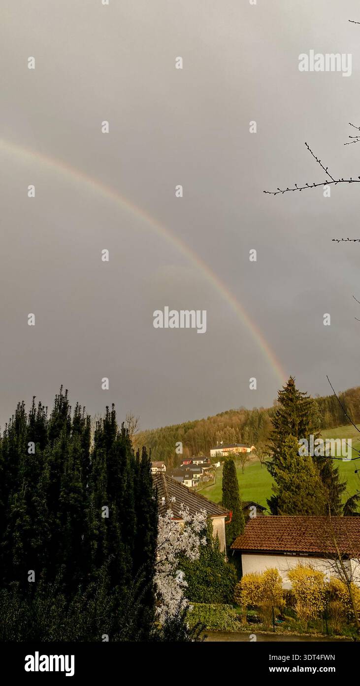 Ein schwacher Regenbogen in einem grauen bewölkten Himmel über Häusern und grünen Bäumen in einem Wohngebiet. Stockfoto