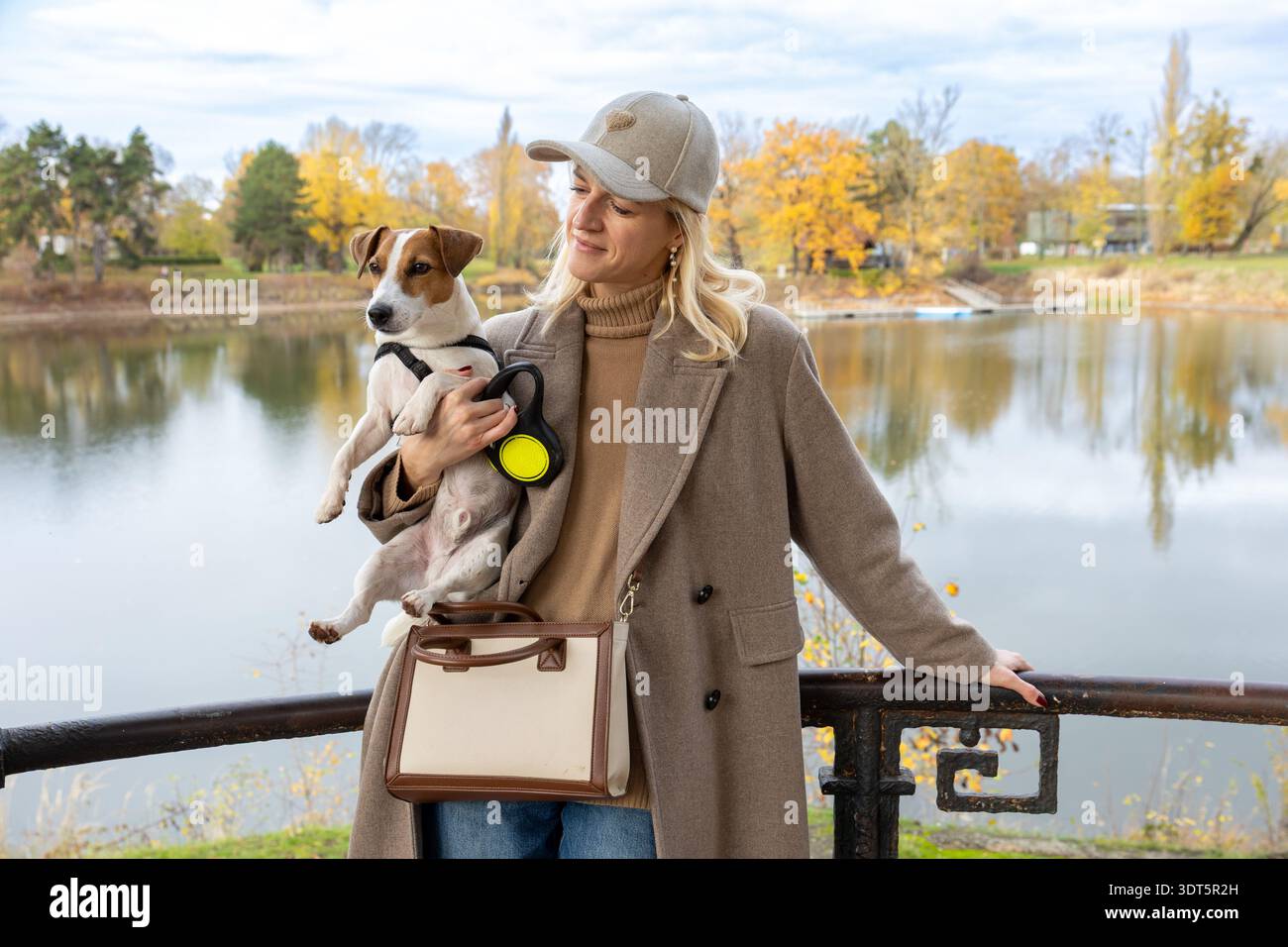 Stilvolle Frau, Die Einen Kleinen Hund In Der Nähe Des Sees Hält, Während Herbst Familienleben Porträt Stockfoto