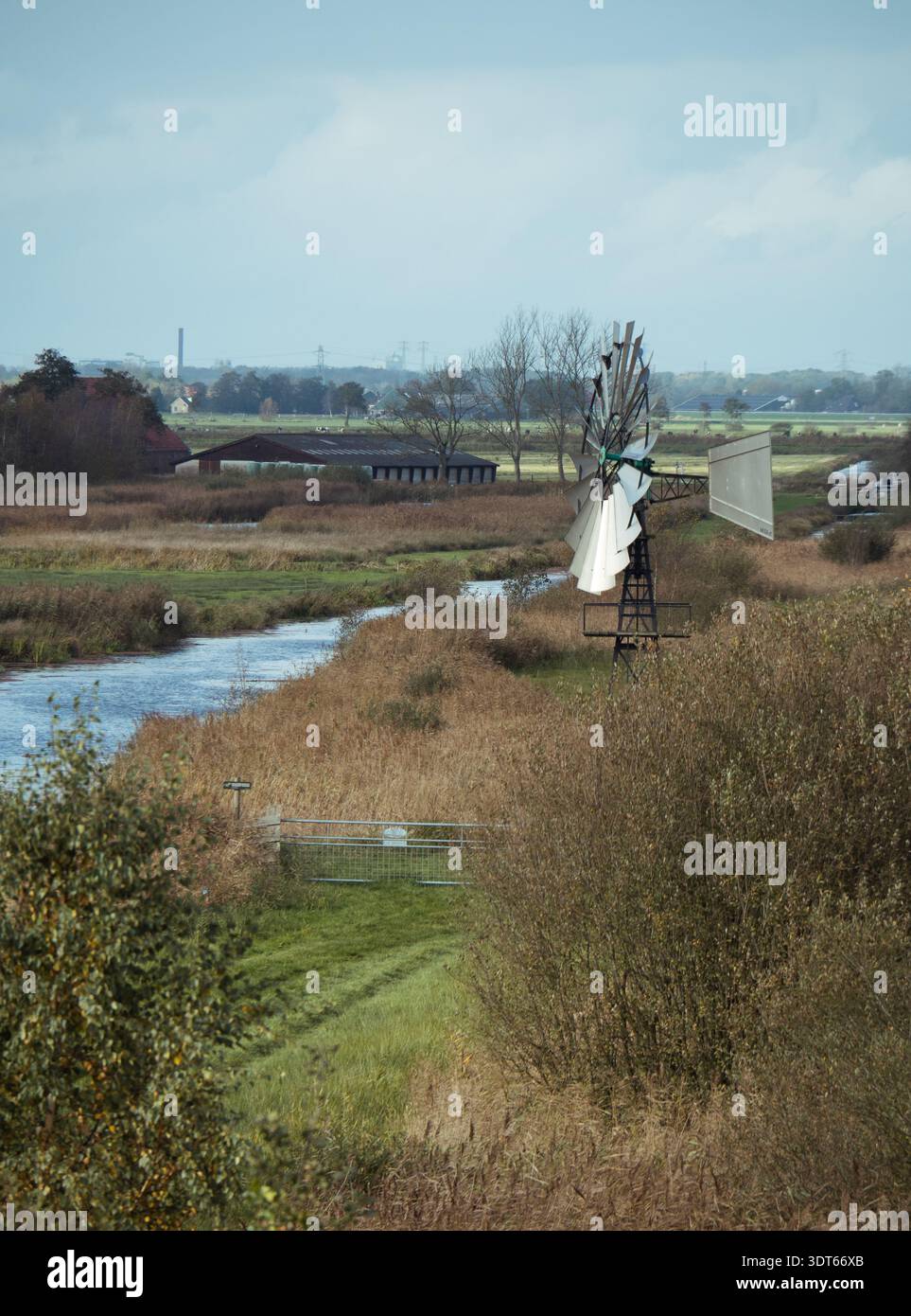 De ALDE Feanen, Niederlande - 27. Oktober 2024: Windmühle steht an einem ruhigen Kanal zwischen Herbstschilf und ländlichen Feldern unter bewölktem Himmel. Stockfoto