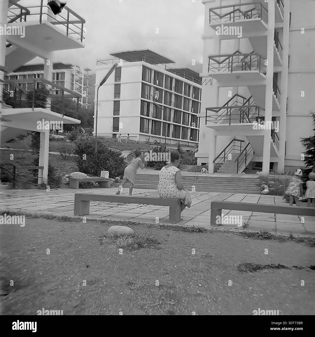 Ein historisches Schwarzweiß-Archivfoto aus den 1960er Jahren, das die beeindruckende modernistische Architektur eines mehrstöckigen Schlafsaals im legendären All-Union Artek Pionierlager in Gurzuf auf der Krim feststellt. Die Szene zeigt Frauen und Kinder, die auf einer langen Bank in einem gepflasterten Innenhof ruhen, mit der innovativen Struktur mit Außentreppen und zahlreichen Balkonen im Hintergrund. Umgeben von üppigem Grün und der malerischen Landschaft der Krim veranschaulicht dieses Bild die friedliche Atmosphäre organisierter Sommerferien in der UdSSR. Stockfoto