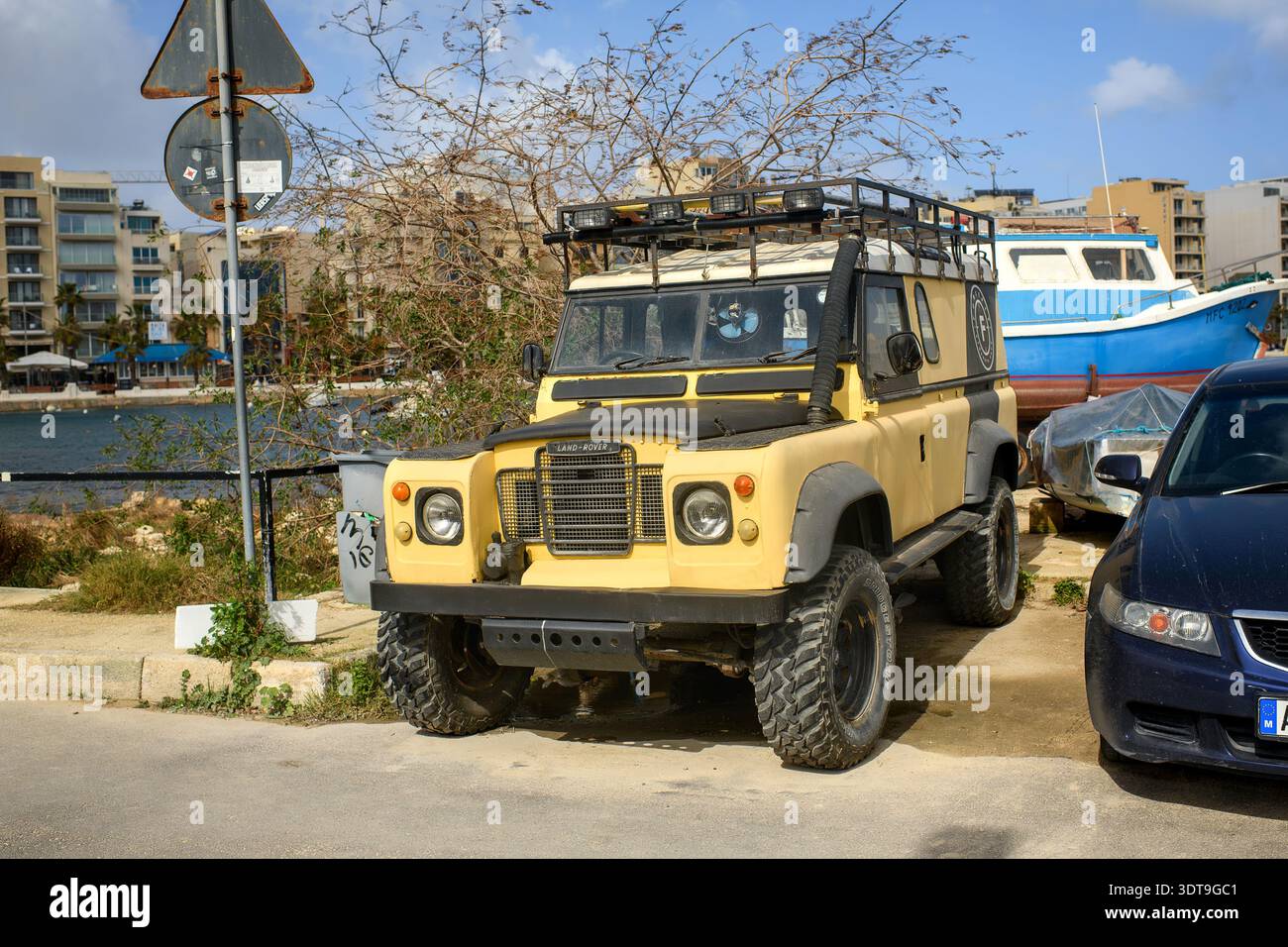 Gzira, Malta - 17. Februar 2026: Ein gelber, gelber Land Rover mit Dachträger und Schnorchel parkt auf der Promenade in der Nähe des Meeres mit Booten. Stockfoto
