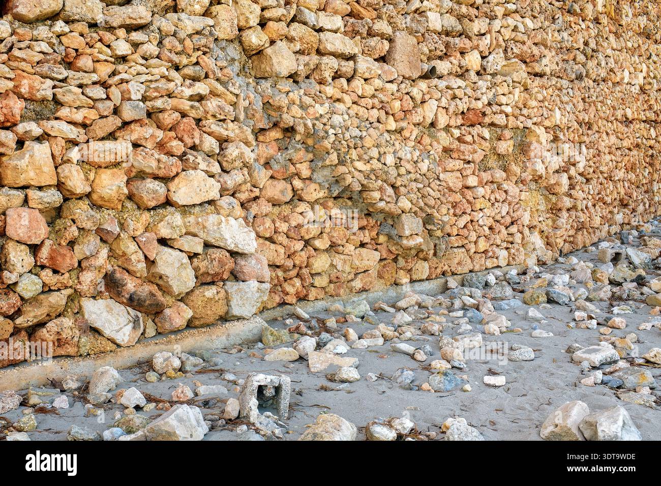 Hochauflösende Aufnahme einer alten rustikalen Steinmauer aus natürlichem Felsen, der auf einer Sandoberfläche mit verstreuten Kieselsteinen und organischen Abfällen steht. Stockfoto
