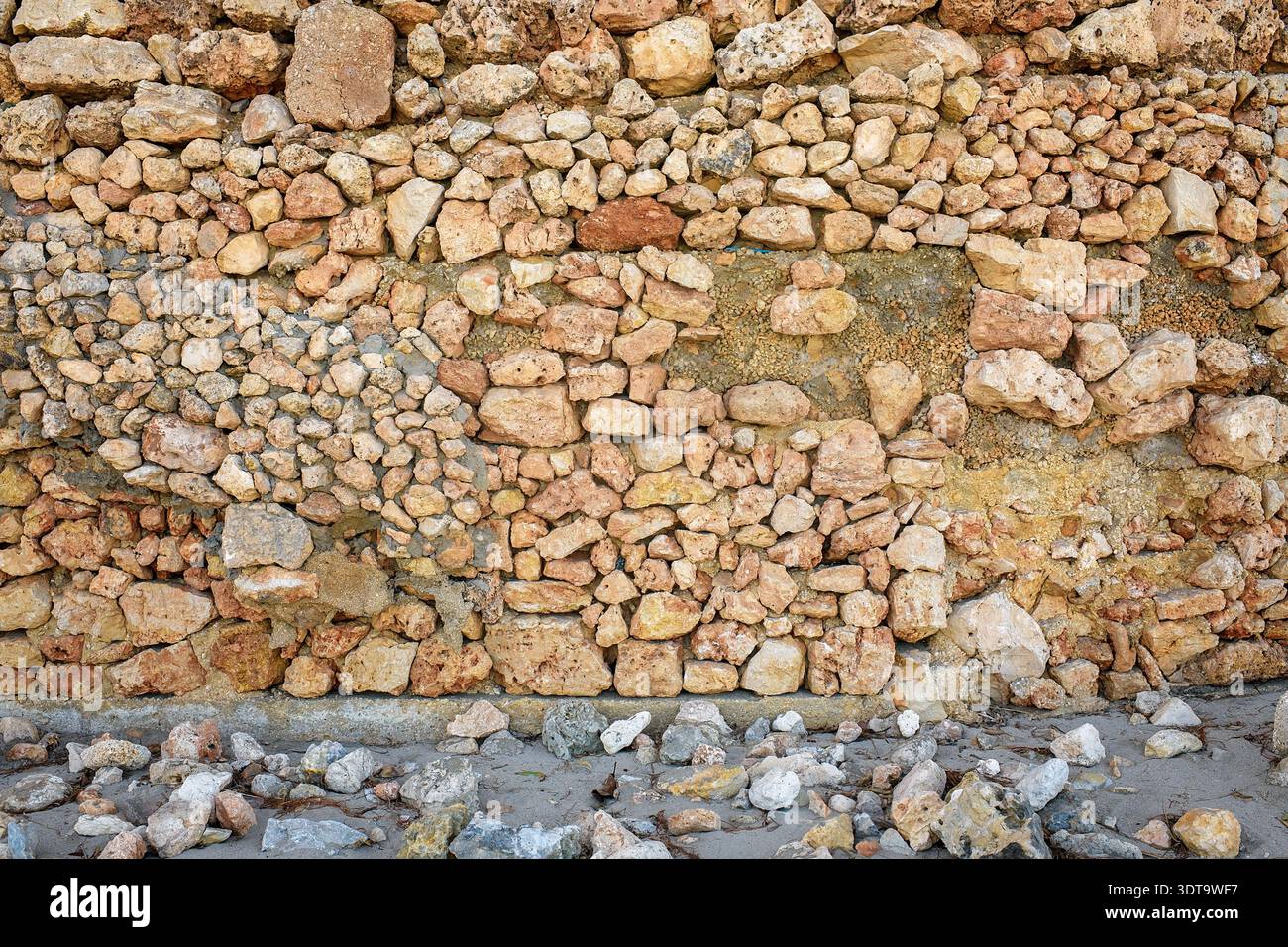 Hochauflösende Aufnahme einer alten rustikalen Steinmauer aus natürlichem Felsen, der auf einer Sandoberfläche mit verstreuten Kieselsteinen und organischen Abfällen steht. Stockfoto