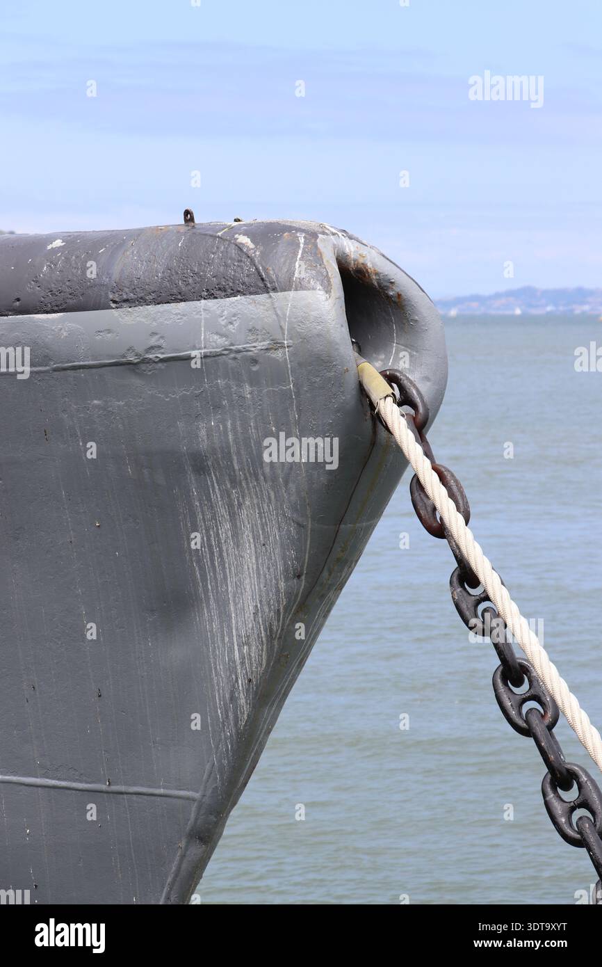 Nahaufnahme eines historischen U-Boot-Bugs, der im Hafen verankert ist und einen verwitterten grauen Stahlrumpf, eine schwere Ankerkette und ein Seil gegen einen ruhigen Küstenstreifen zeigt Stockfoto