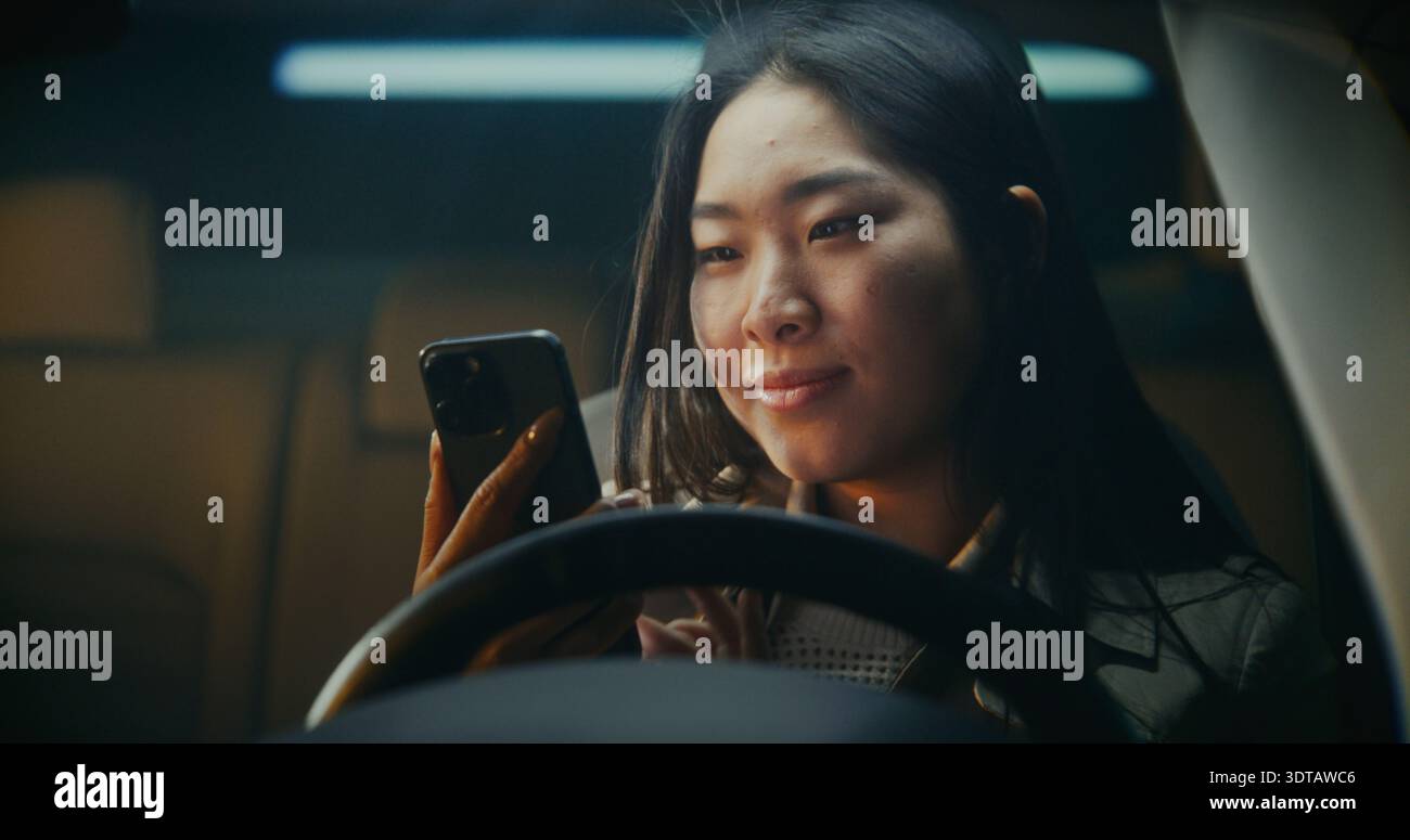 Die junge Frau steigt in das Auto und nimmt das Smartphone auf, öffnet es mit Gesichtserkennung. Ausdruck wird in warmes Lächeln gemildert, wenn neue Nachricht auf dem Bildschirm angezeigt wird. Moderner Digitaler Lifestyle, Alltägliche Konnektivität. Stockfoto