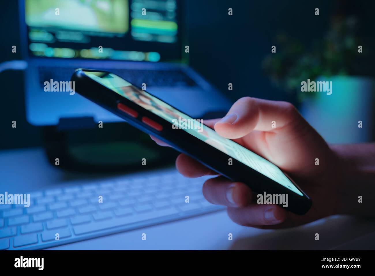 Nahaufnahme von Inhalten auf einem Mobiltelefon vor einem Schreibtisch mit Laptop in der Nacht. Digitales Leben, Inhaltskonsum, sucht nach sozialen Medien und ständige Online-Interaktion. Stockfoto