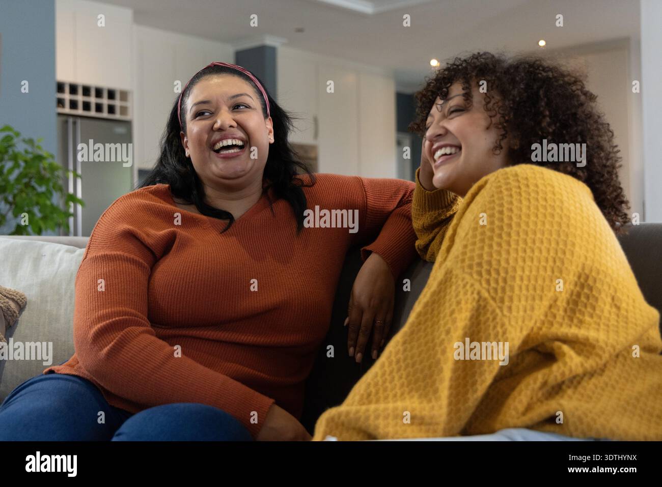 Verschiedene Freundinnen lachen und sitzen zu Hause auf der Couch, tragen Rost- und Senfpullover Stockfoto
