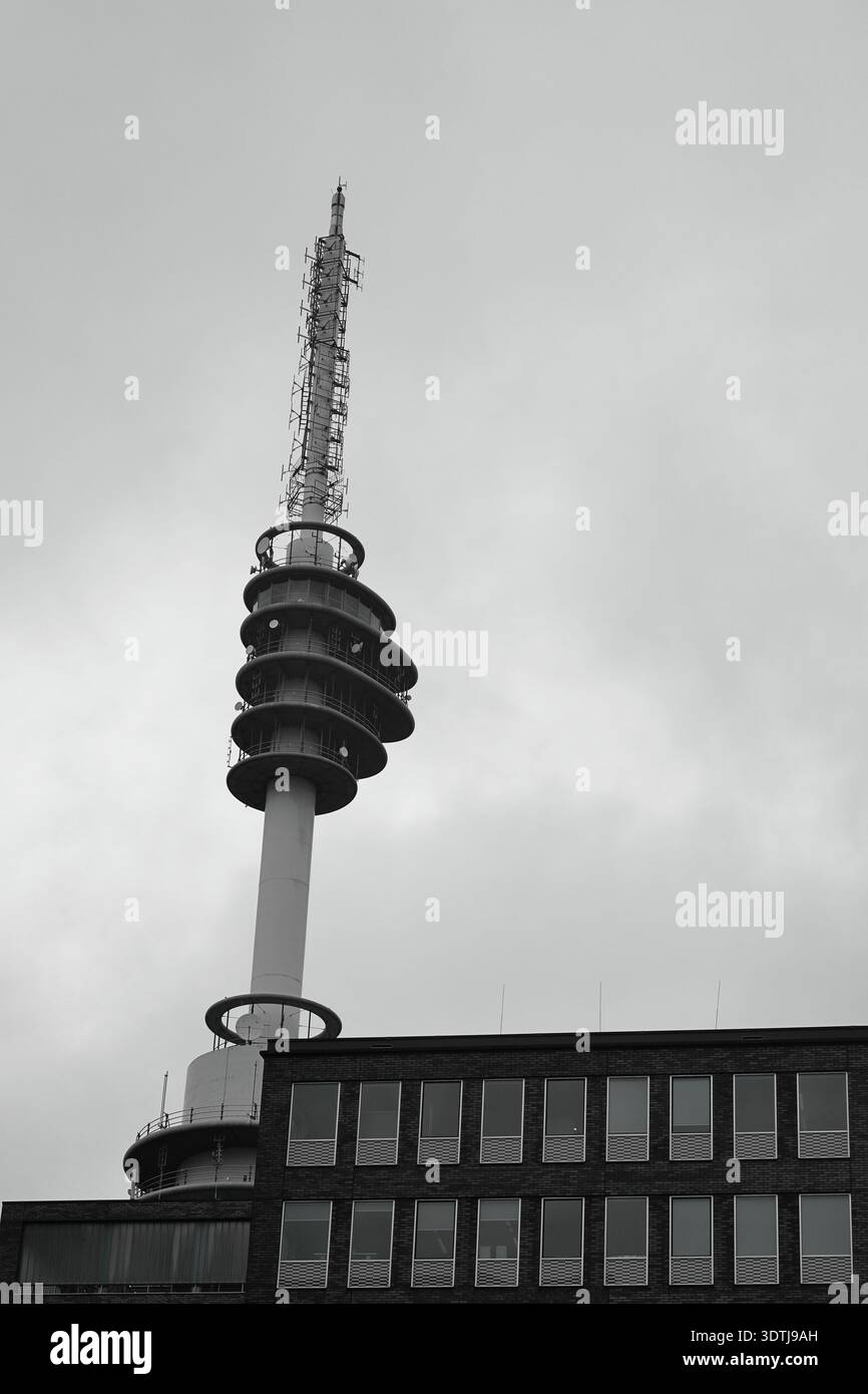 Schwarz-weiß-Blick auf einen Kommunikationsturm über Backsteinbürogebäuden in Amsterdam, Niederlande, unter einer dramatischen, bewölkten Winterskyline. Stockfoto