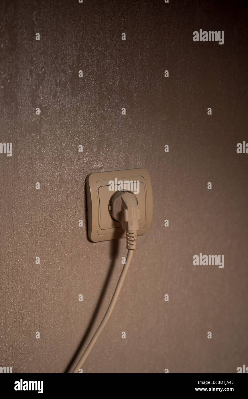 Ein beigefarbener Stecker sitzt in einer europäischen Einbausteckdose an einer taupefarbenen Wand in Innenräumen unter warmem, direktionalem Licht, das einen weichen Kabelschatten in einem minimalistischen C erzeugt Stockfoto
