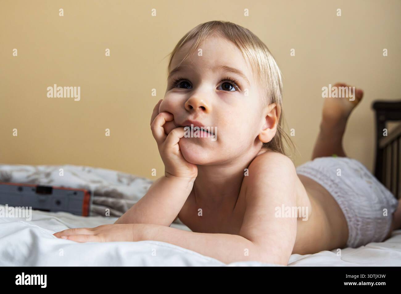 Nachdenkliches blondes Kleinkind, das auf weißem Bett liegt und aufblickt, Finger in Mundnähe Stockfoto