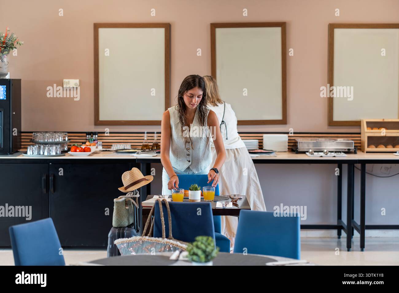 Eine Frau steht an einem Tisch im Essbereich des Hotels und arrangiert Gläser Saft. Die moderne und einladende Umgebung lässt eine friedliche Morgenroutine erwarten Stockfoto