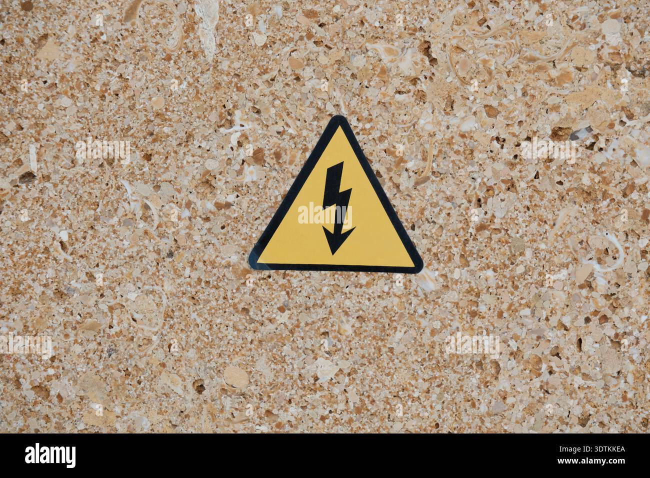 Gelbes Warnschild mit Blitzsymbol an strukturierter beiger Wand. Das Schild weist auf elektrische Gefahren und Sicherheitsvorkehrungen hin. Stockfoto