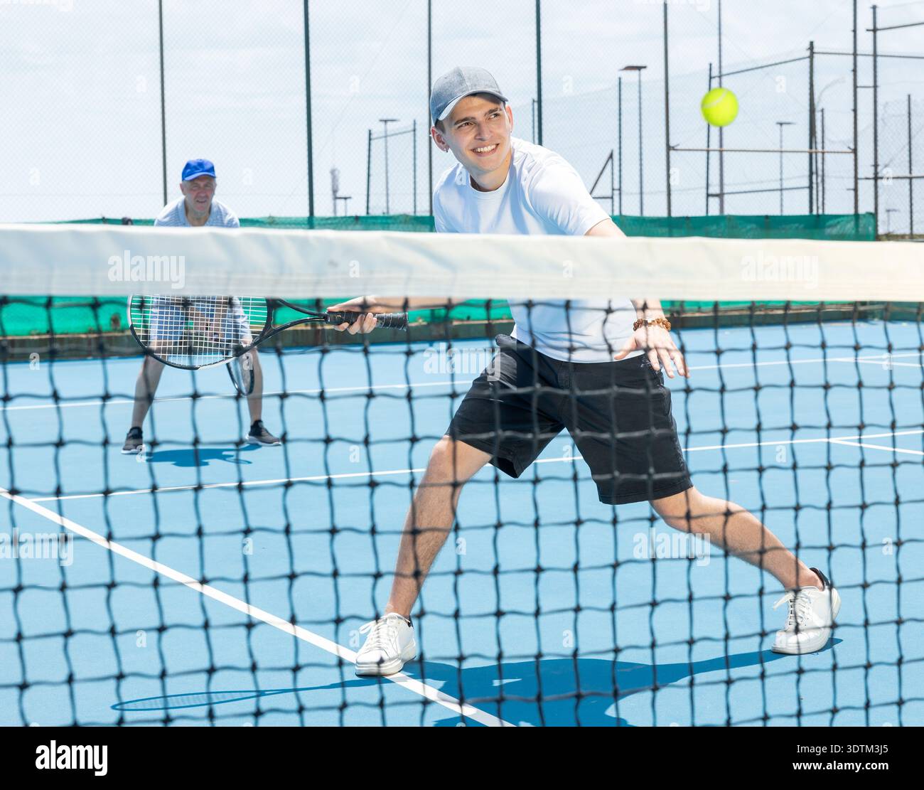 Porträt eines jungen Mannes, der im Sommer auf dem offenen Platz Tennis spielt, und Schläger schwingt, um den Ball über das Netz zurückzubringen. Sportler bereit für Volley Stockfoto