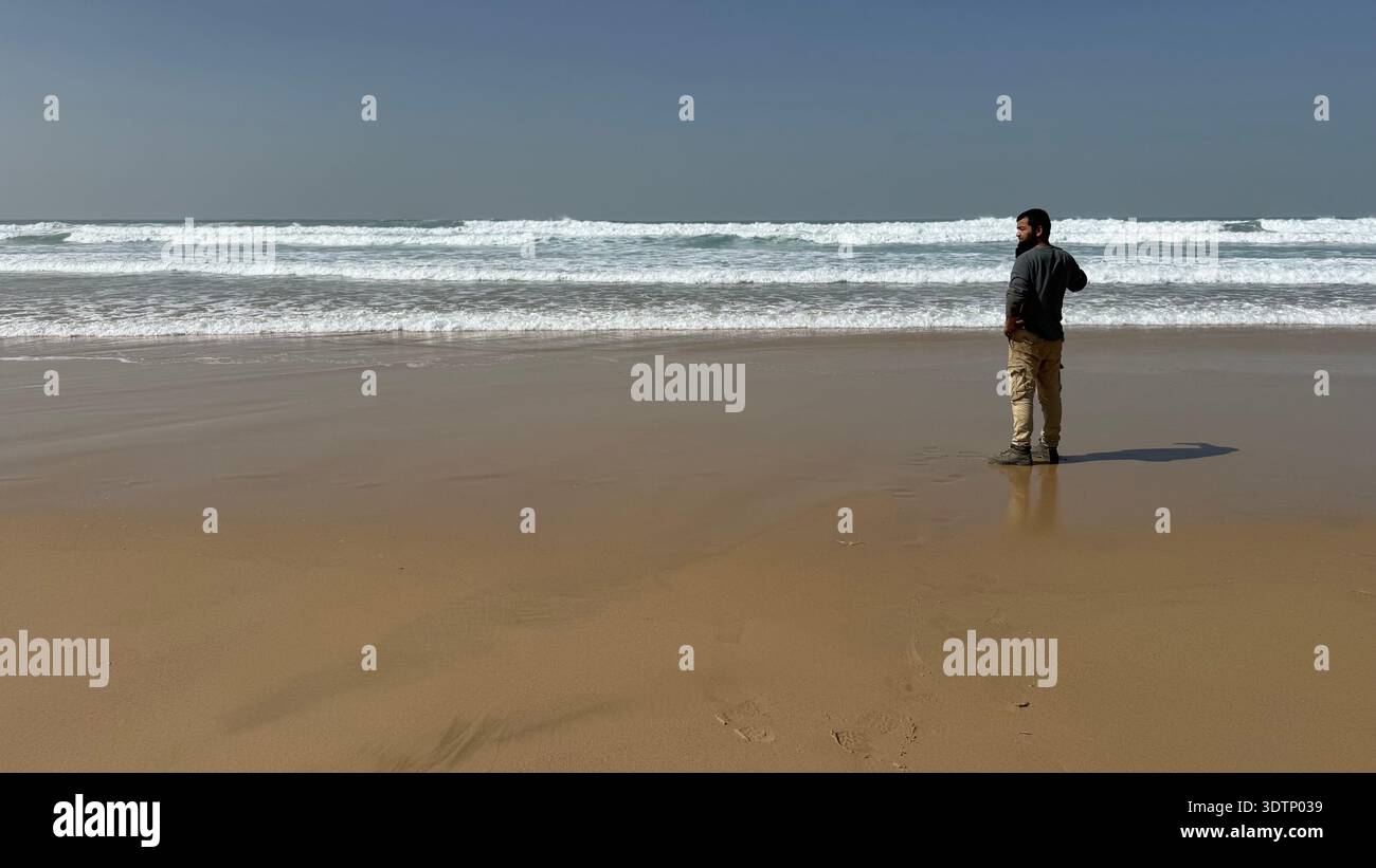 Seitenprofil eines Mannes, der auf einem riesigen, goldenen Sandstrand steht und auf die weißen Wellen des Ozeans unter einem klaren blauen Himmel blickt. Das Konzept von Einsamkeit und Frieden - Smartphone-aufgenommenes Stockfoto