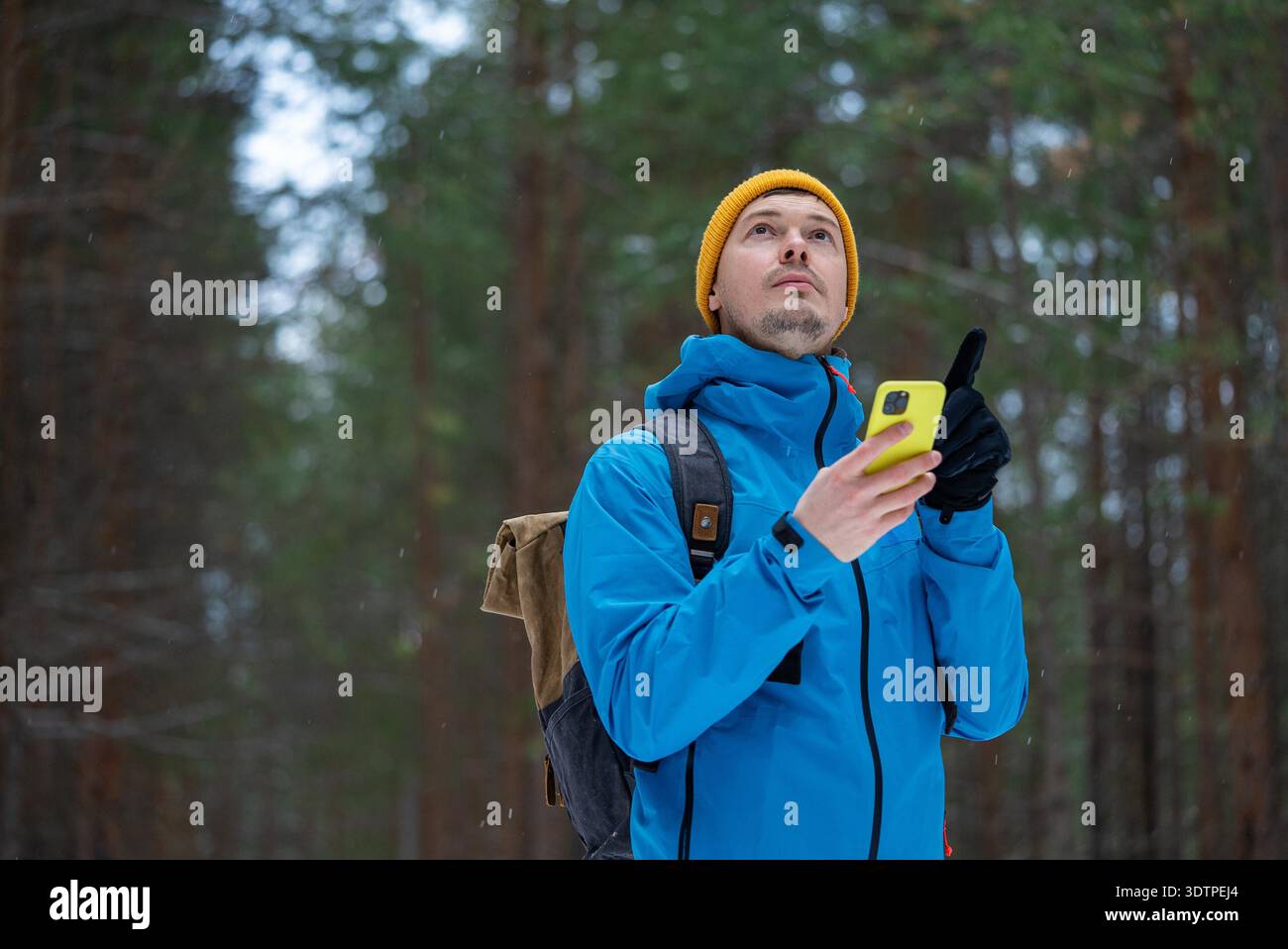 Mann in Winterkleidung, der Smartphone für die Navigation nutzt, sich verloren fühlt und in einem verschneiten Wald nach der Richtung sucht, symbolisiert Herausforderung, Erkundung und moderne Technologie-Abhängigkeit || Model veröffentlicht Stockfoto