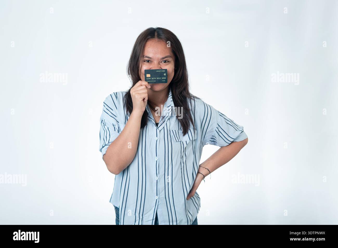 Eine indonesische Frau in einem blau gestreiften Hemd, die eine hochwertige schwarze Kreditkarte vor dem Gesicht hält und ihren Mund bedeckt. Lächelnde Augen und selbstbewusste Pose Stockfoto