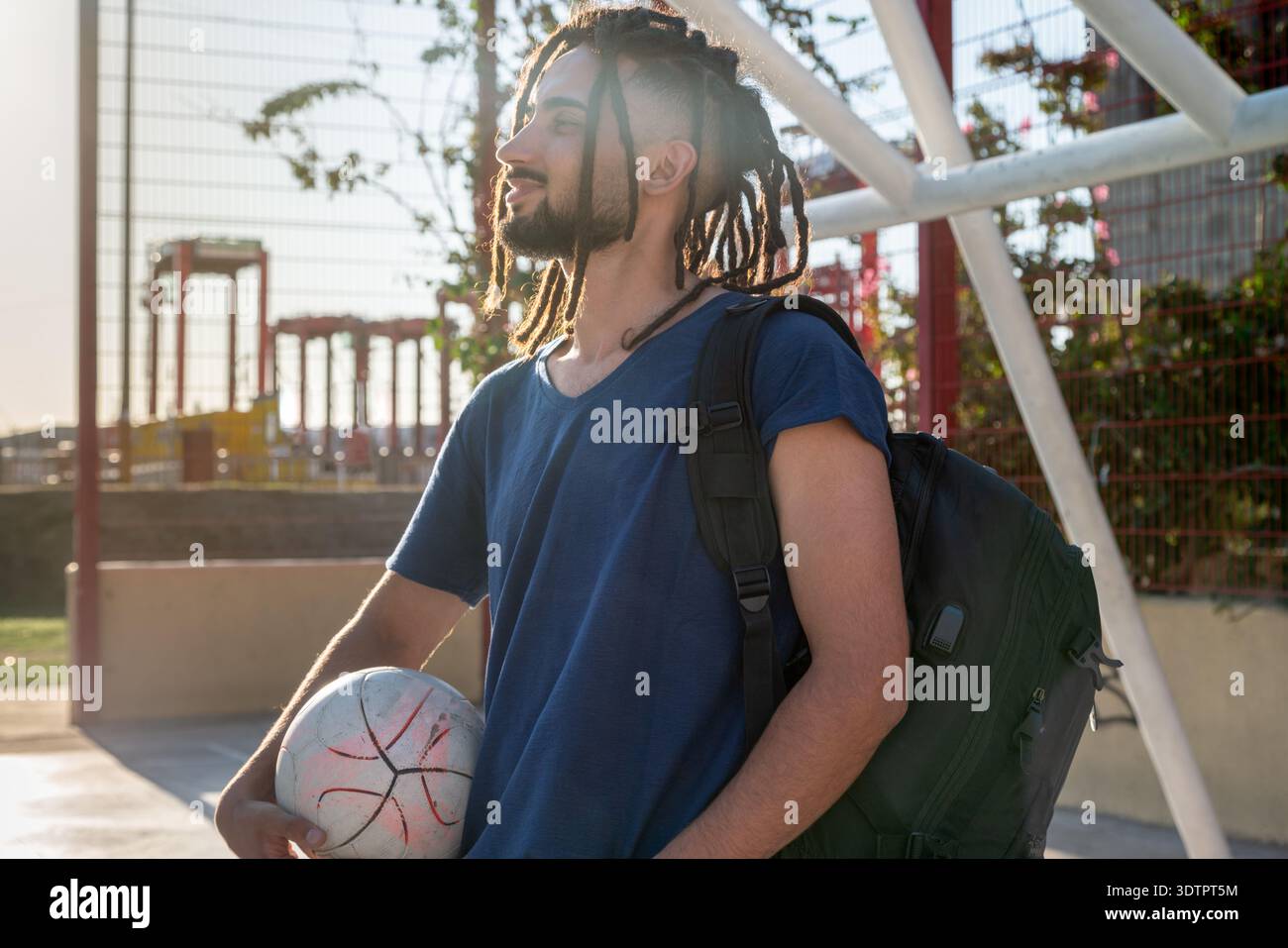 Junger Mann mit Dreadlocks, der einen Fußball auf einem Straßengarten hält und wegschaut Stockfoto