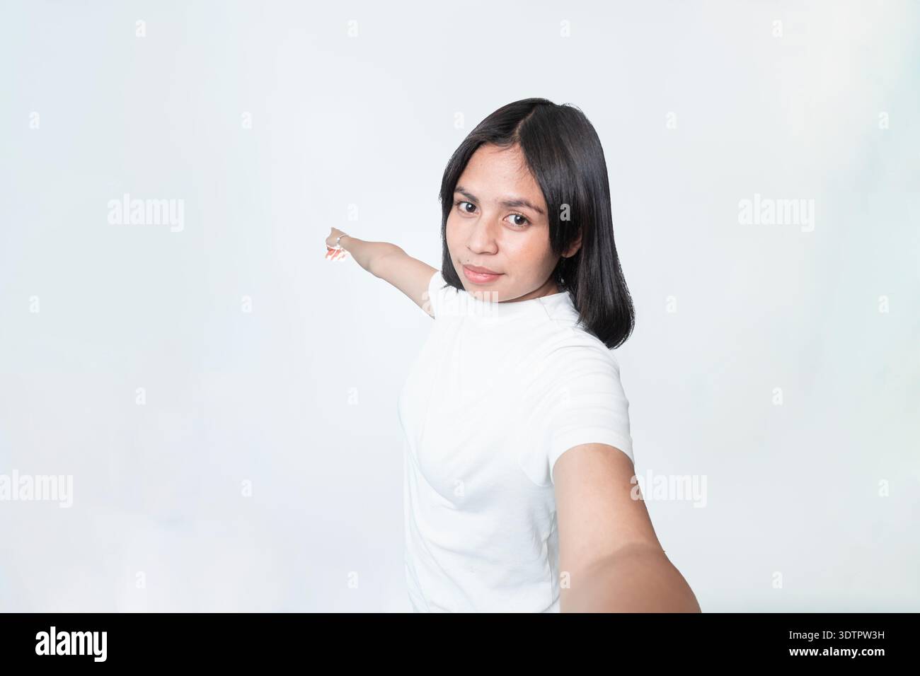 Eine junge indonesische Frau in einem weißen T-Shirt, die mit dem Finger nach hinten zeigt, während sie in die Kamera lächelt. Isoliert auf weißem Hintergrund mit Kopierraum. Stockfoto