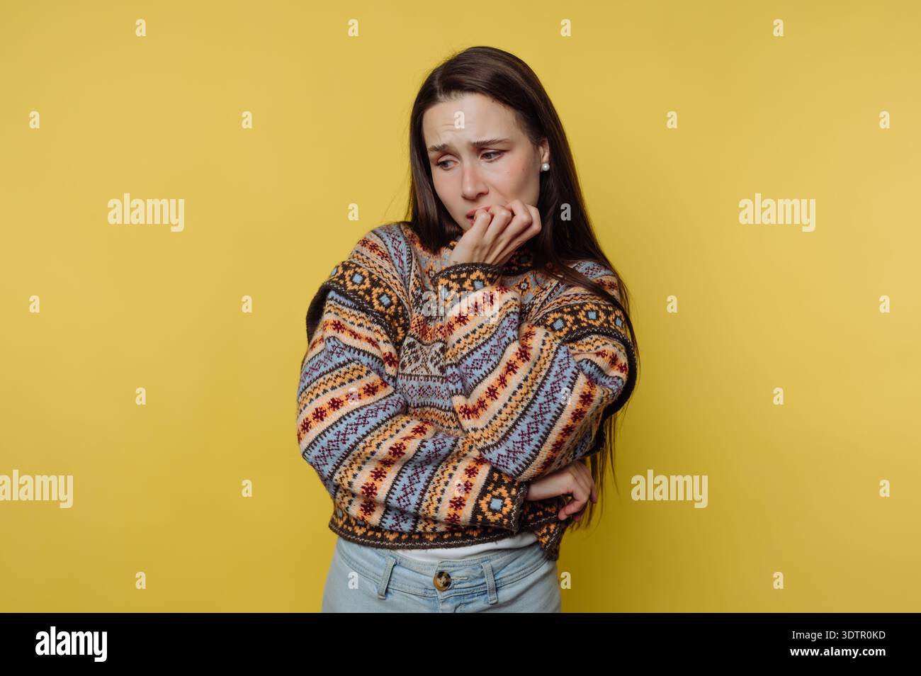 Ein Mädchen in einem farbenfrohen Strickpullover steht mit überkreuzten Armen, verloren in Gedanken. Stockfoto