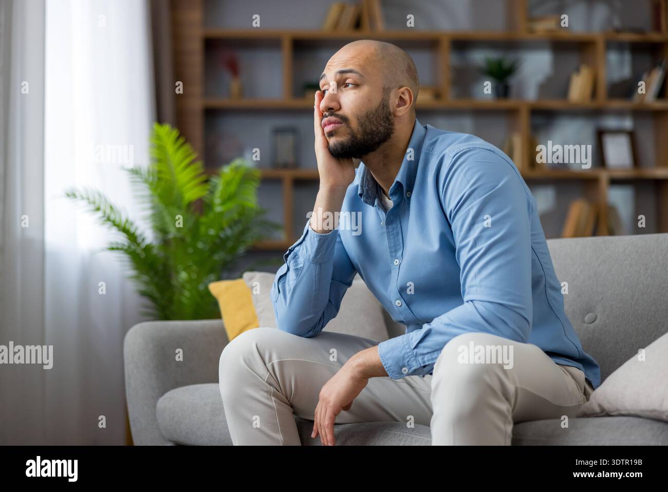 Nachdenklicher Glatzkopf, der allein auf einem grauen Sofa in einem modernen Wohnzimmer sitzt und wegblickt, während er nachdenkt, reflektiert und Stress oder Enttäuschung erlebt Stockfoto