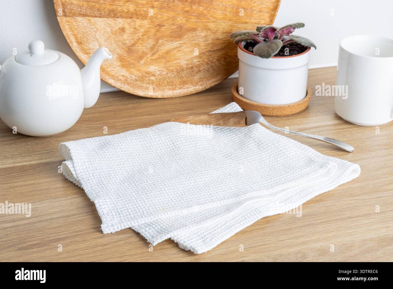 Eine ordentlich gestaltete Küchenszene mit einem weißen Teeset, einem Holzschneidebrett und einer Topfpflanze auf einem Holztisch. Stockfoto