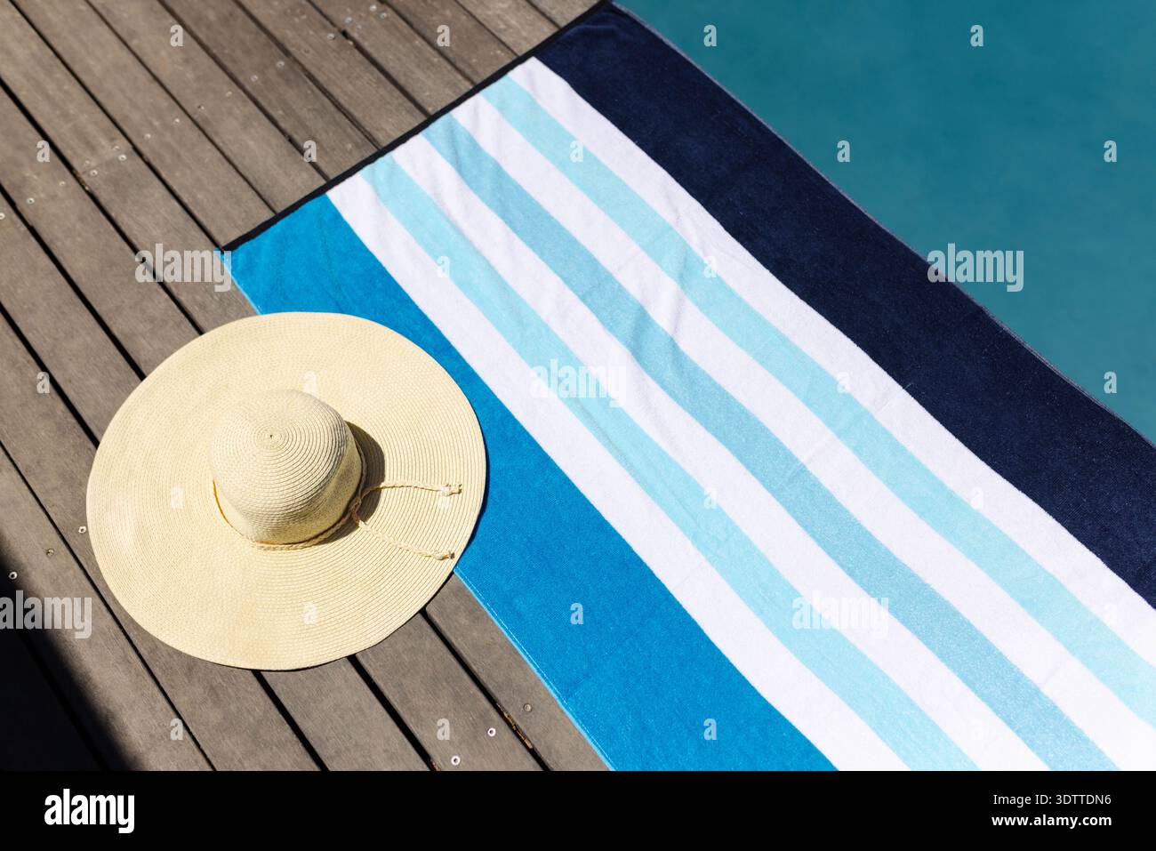 Gestreiftes Strandtuch liegt flach auf einer sonnendurchfluteten Holzterrasse, mit Strohhut in der Nähe des Pools und Schatten Stockfoto