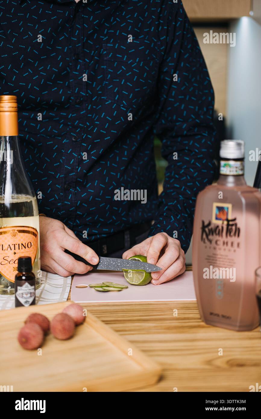Person schneidet Limetten auf Schneidebrett, umgeben von Platino-Wein und Kwai-Feh-Likörflaschen. Barkeeper, Craft Cocktail Bewegung, DIY Drinks Stockfoto