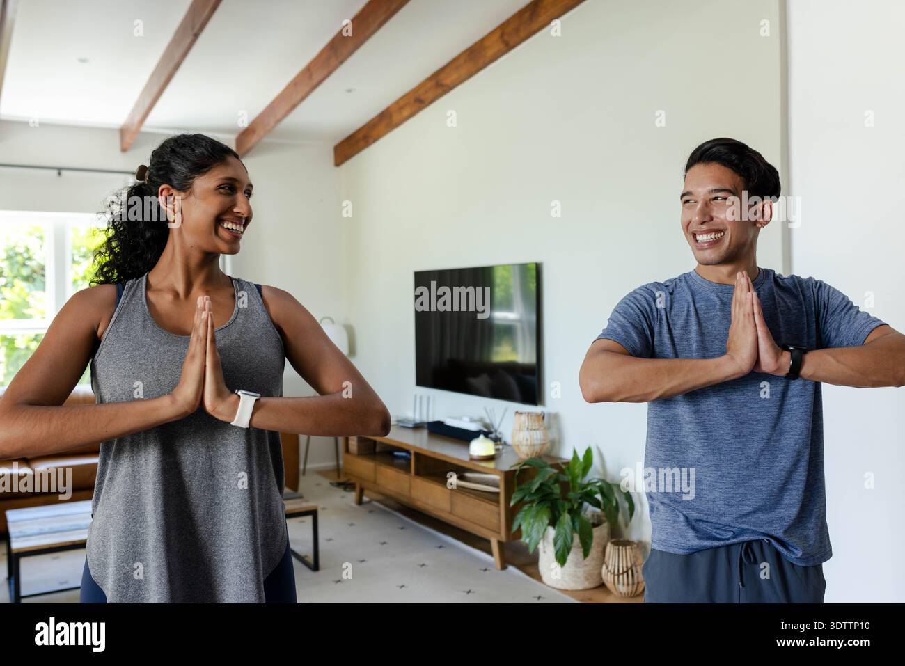 Afrikanisches Paar, das im Wohnzimmer mit Fernseher gepaart Yoga praktiziert, Sportkleidung und Smartwatches trägt Stockfoto