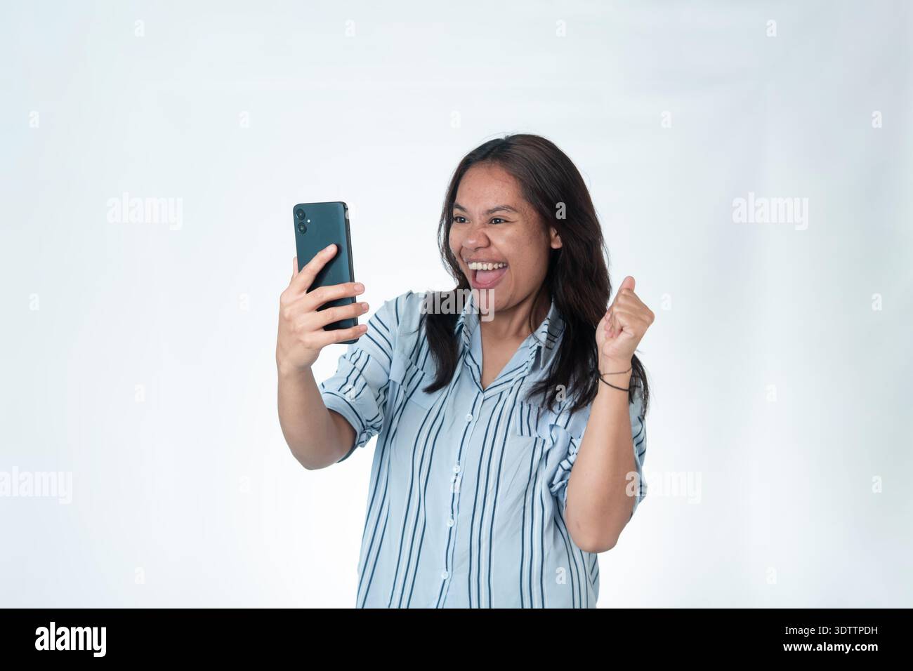 Fröhliche indonesische Frau in einem blau gestreiften Hemd, hält ein Smartphone und ballt ihre Faust in einer gewinnenden Geste. Sie hat ein großes fröhliches Lächeln und Klo Stockfoto