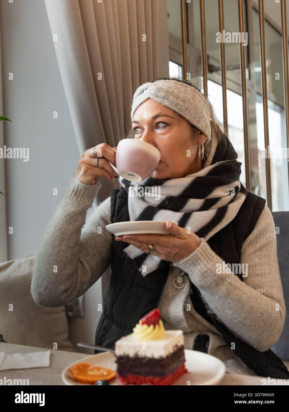 Frau trinkt Kaffee, während sie an einem Café-Tisch mit Dessertkuchen in einem modernen, gemütlichen Café-Interieur sitzt. Stockfoto