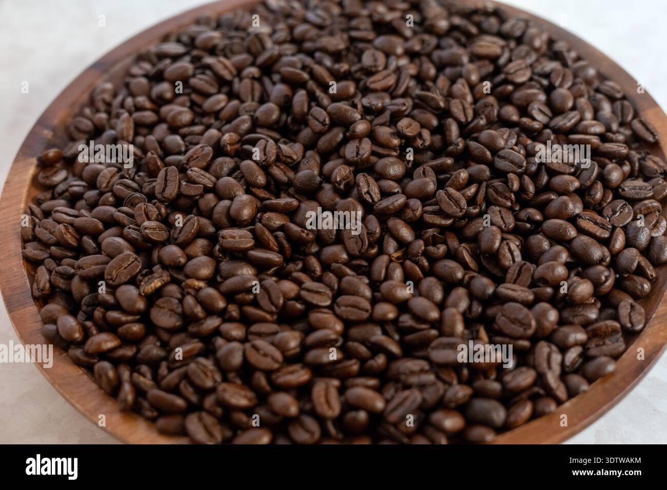 Eine professionelle Aufnahme dunkel gerösteter Kaffeebohnen, die eine runde Holzschale füllen, die auf einer hellen weißen Marmoroberfläche platziert ist und natura unterstreicht Stockfoto