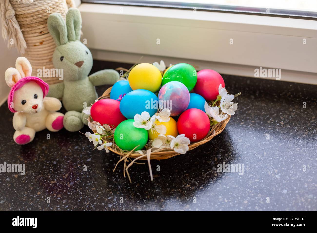 Bunte Ostereier in einem Korb neben kleinen Plüschhasen auf dunkler Oberfläche. Urlaubsarrangements, Frühlingsdekor, festliche Stimmung. Ostern. Stockfoto