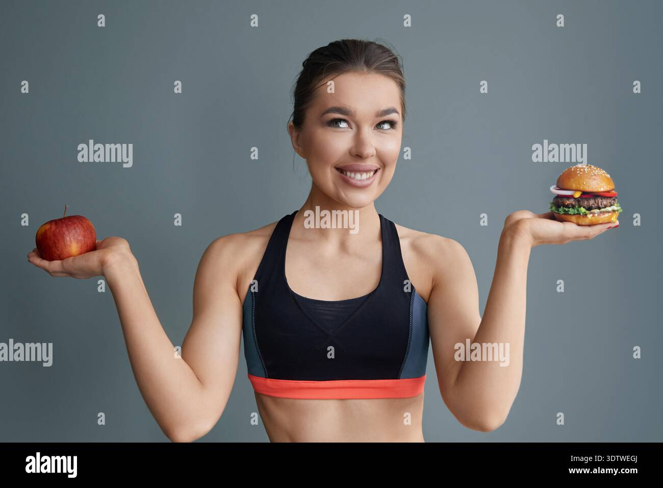 Eine Frau steht mit einem Apfel in der linken Hand und einem Burger in der rechten und lächelt selbstbewusst. Sie trägt ein sportliches Outfit und ist gegen einen einfachen bac gestellt Stockfoto