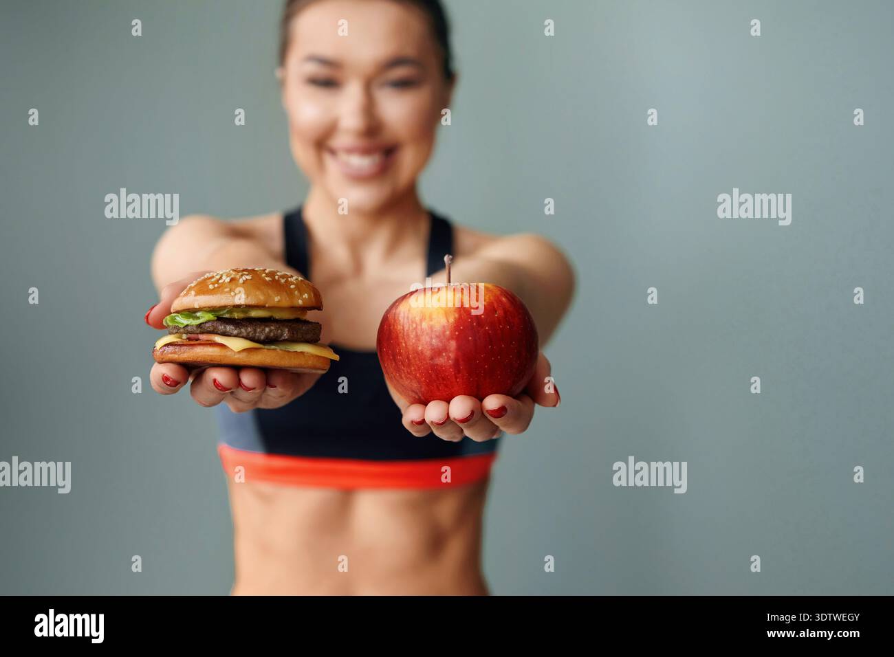 Eine Frau lächelt weit und hält einen Apfel in der linken Hand und einen Burger in der rechten Hand. Sie trägt Sportkleidung und steht vor einem einfachen B Stockfoto