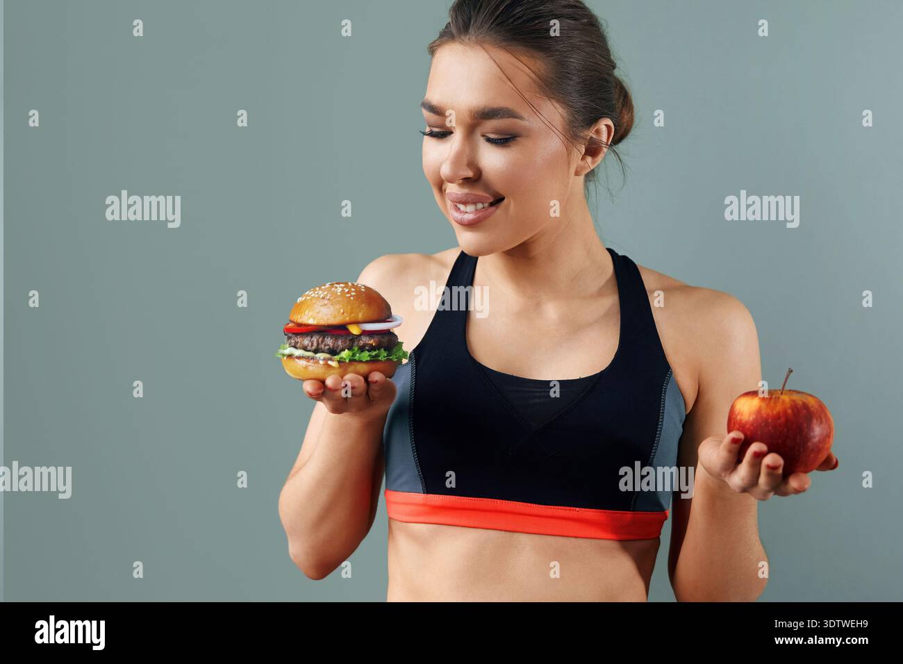 Eine junge Frau in Sportkleidung steht vor grauem Hintergrund, hält einen Apfel in der einen Hand und einen Burger in der anderen und denkt über ihre Essensauswahl nach Stockfoto