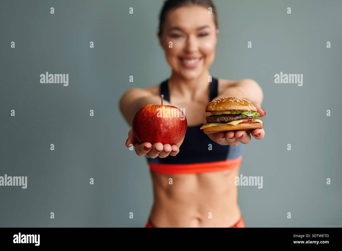 Eine Frau lächelt weit und hält einen Apfel in der linken Hand und einen Burger in der rechten Hand. Sie trägt Sportkleidung und steht vor einem einfachen B Stockfoto