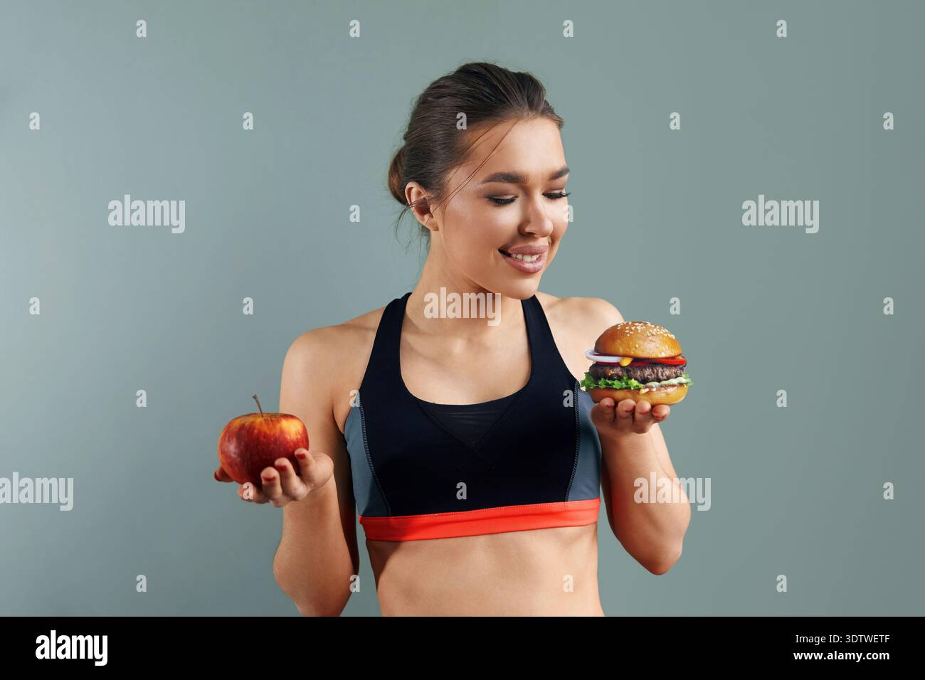 Eine junge Frau in Sportkleidung steht vor grauem Hintergrund, hält einen Apfel in der einen Hand und einen Burger in der anderen und denkt über ihre Essensauswahl nach Stockfoto
